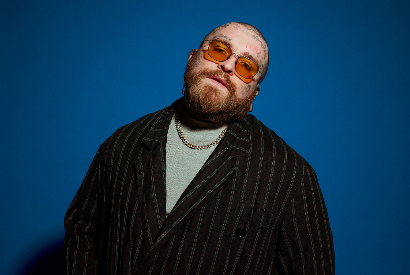 man with face tattoos, a ginger beard and sunglasses, standing against a blue background